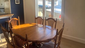 Un coin repas avec une table et des chaises en bois, un luminaire suspendu, un espace cuisine équipé d’appareils électroménagers et une fenêtre donnant sur l’extérieur.