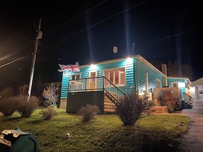 A small, single-story house with a porch, a flag, and a well-lit front yard.