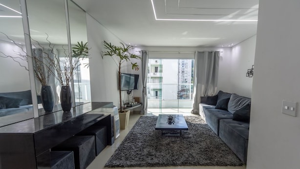 A modern living room with a sofa, a coffee table, a TV, and a large window. - Apto com Churrasqueira e Vaga a 200m da Praia (Guarujá)