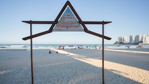 A beach sign with a triangular structure and a message in Portuguese. - Apto com Churrasqueira e Vaga a 200m da Praia (Guarujá)