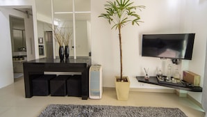 A modern living room with a flat-screen TV, a dining area with a black table and chairs, a potted plant, and a kitchen in the background. - Apto com Churrasqueira e Vaga a 200m da Praia (Guarujá)