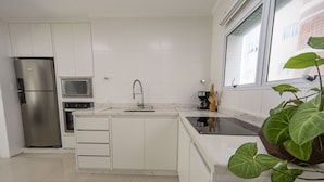 A modern kitchen with white cabinetry, a stainless steel refrigerator, a microwave, a sink with a faucet, and a window with a view of buildings. - Apto com Churrasqueira e Vaga a 200m da Praia (Guarujá)