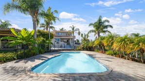 A swimming pool with a paved deck, surrounded by palm trees and a building with a tiled roof in the background.