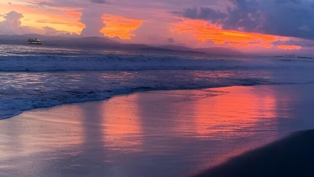Une plage avec un navire à l ’horizon, des vagues et un coucher de soleil.