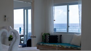 Ett rum med en säng, ett fönster med utsikt över havet, ett litet bord med en växt och en dörr som leder ut.