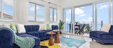 A modern living room with a blue sofa, a coffee table, and a view of the sea through large windows.