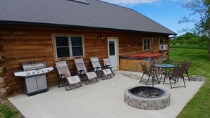 Una cabaña de madera con una parrilla, sillas de lounge, una fogata y una mesa con sillas.