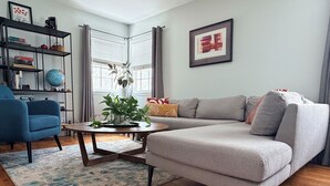 Een woonkamer met een grijze bank, een blauwe fauteuil, een salontafel, een boekenkast en een raam met gordijnen.
