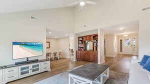 Un vaste salon doté d’un téléviseur à écran plat, d’un coin repas, d’une cuisine avec des meubles en bois et d’une vue sur la plage par la fenêtre.