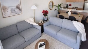 Ein modernes Wohnzimmer mit grauen Sofas, einem Couchtisch, einem kleinen Beistelltisch mit Lampe, einem gerahmten Bild an der Wand und einer Küche im Hintergrund.