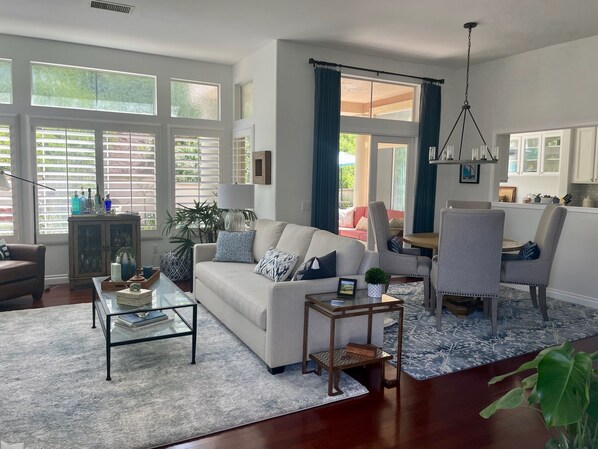 Un salón espacioso con sofá, mesa de centro y sillas, ventanas grandes con persianas y vistas al exterior.