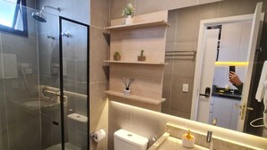 Et moderne badeværelse med en glasbrusekabine, toilet, vask med granitbordplade og træhylder over toilettet.