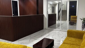 A reception area with a wooden reception desk, a yellow sofa, a dark wooden coffee table, and a ceiling fan. - Hotel NB aeropuerto (Puerto Vallarta)