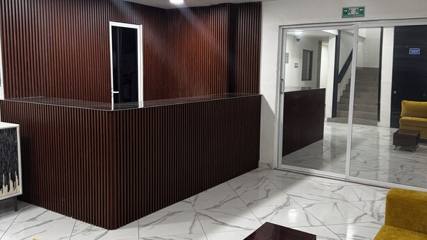 A reception area with a wooden reception desk, yellow sofas, a marble floor, and a ceiling fan. - Hotel NB aeropuerto (Puerto Vallarta)