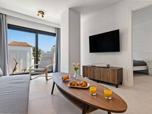 Ein modernes Hotelzimmer mit Balkon, Flachbildfernseher, Esstisch mit Frühstück und einer Couch.
