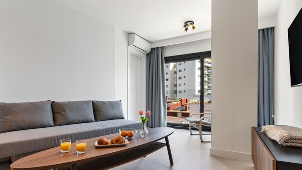 A modern living room with a sofa, a wooden coffee table with breakfast items, a TV, and a view of a building through large windows. - La Joya 1A. New apartment with Garage, 300 m from the beach (Torremolinos)