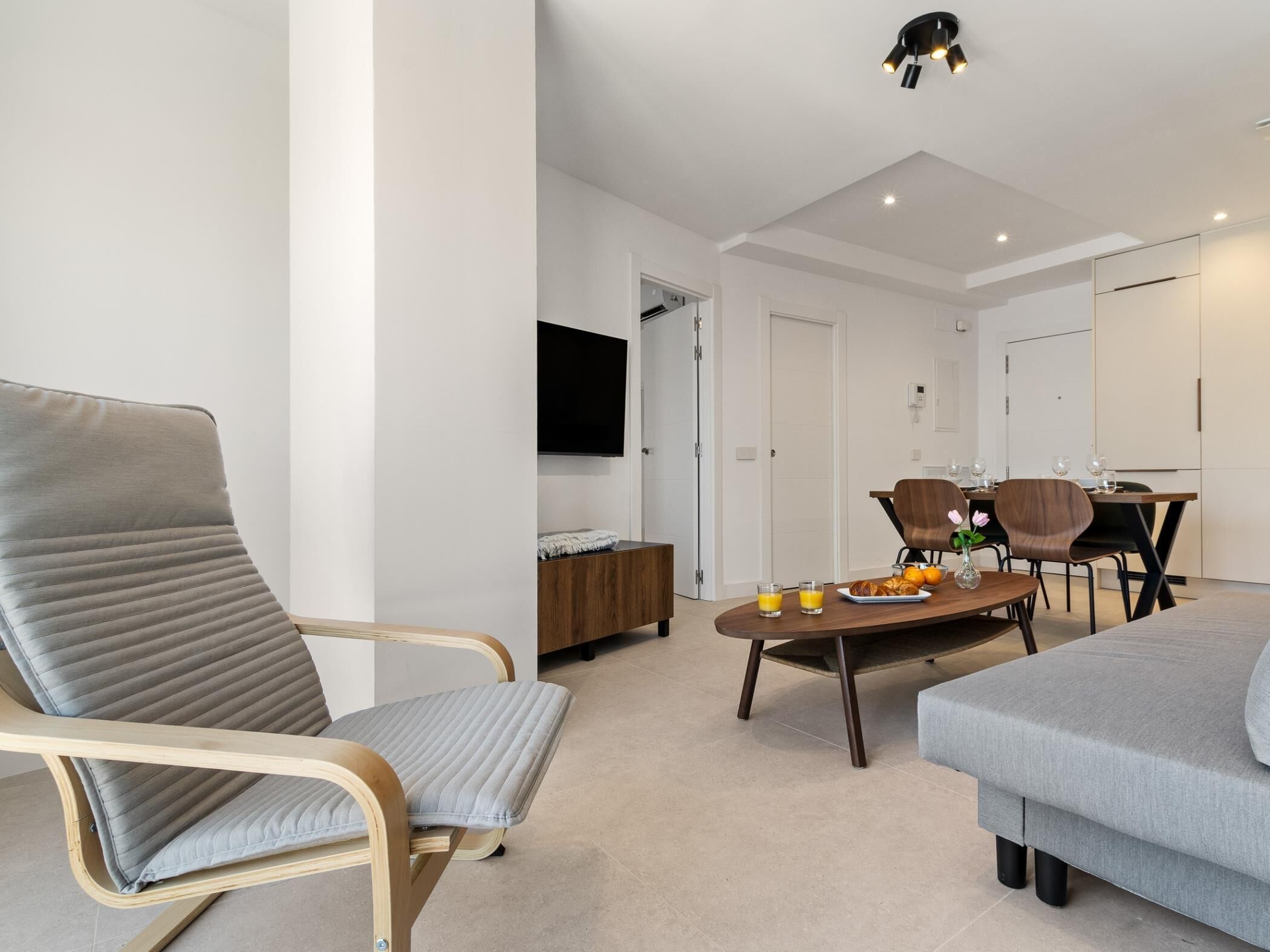 Un salon moderne comprenant un canapé, une table à manger avec des chaises, un téléviseur fixé au mur et un luminaire suspendu au plafond.