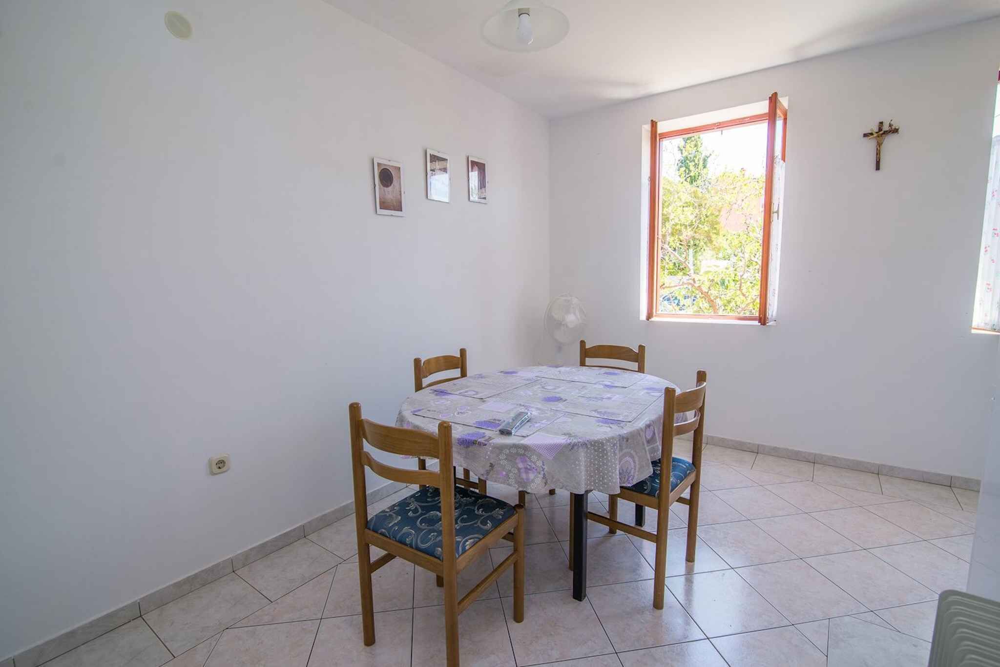 Uma sala de jantar com uma mesa redonda, quatro cadeiras de madeira, uma luminária no teto, uma janela e um detalhe em forma de cruz na parede, além de um chão de azulejos.