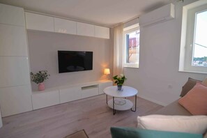 A modern living room with a flat-screen TV mounted on the wall, a small round table with a vase of flowers, a sofa with cushions, and a window with curtains. - Holiday flat Sofia City centre apartment (Mali Lošinj)
