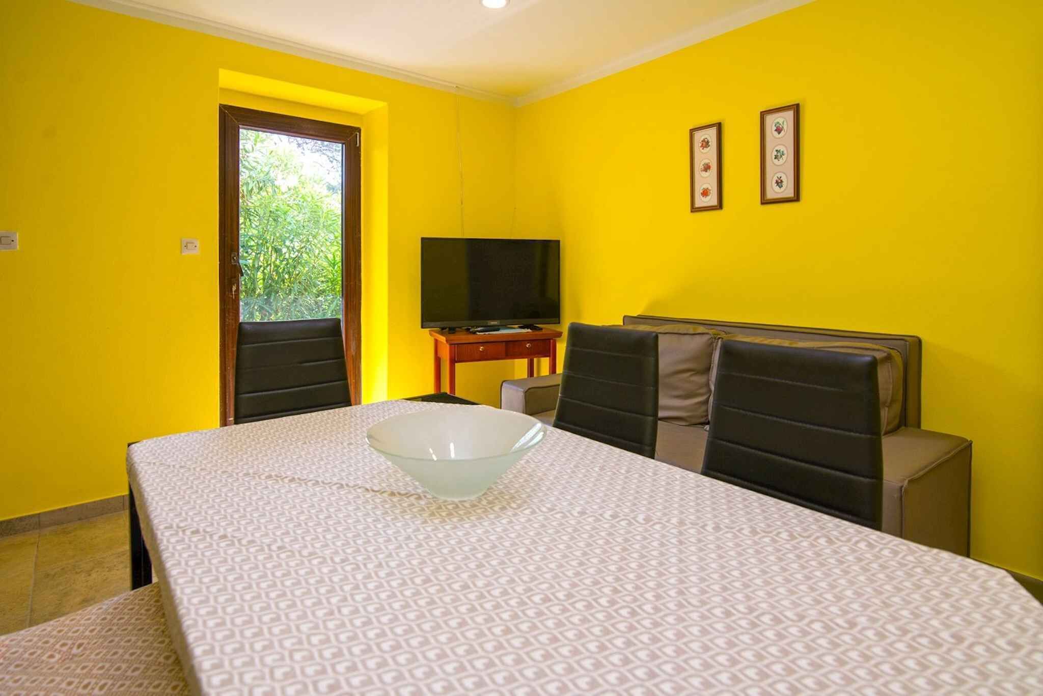 Une pièce aux murs jaunes, un lit recouvert d’une couvre-lit à motifs, un téléviseur posé sur un meuble et une fenêtre donnant sur un paysage verdoyant.