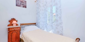 Ein Schlafzimmer mit einem Bett, einem Nachttisch mit Lampe, einer Kommode und einem Fenster mit blumigem Vorhang.
