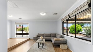 Ein modernes Wohnzimmer mit einem grauen Ecksofa, einem kleinen Tisch mit einer Vase, einem großen Fenster mit Jalousien und Holzboden.