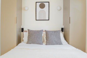 A bed with white bedding, two grey pillows, a framed abstract artwork above the bed, and two wall-mounted light fixtures. - Blueground | Le Marais, view, nr Place des Vosges (Paris)
