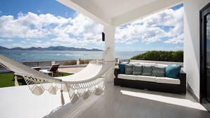 En balkon med hængekøje, sofa med puder og udsigt til hav og bjerge.