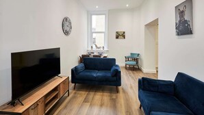 A living room with a flat-screen TV on a wooden stand, two dark blue sofas, a small table, and a window with blinds. - Host Stay The Stables Apartment 1 (Saltburn-by-the-Sea)