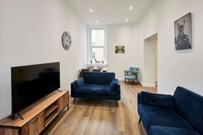 A living room with a flat-screen TV on a wooden stand, two dark blue sofas, a small table, and a window with blinds. - Host Stay The Stables Apartment 1 (Saltburn-by-the-Sea)