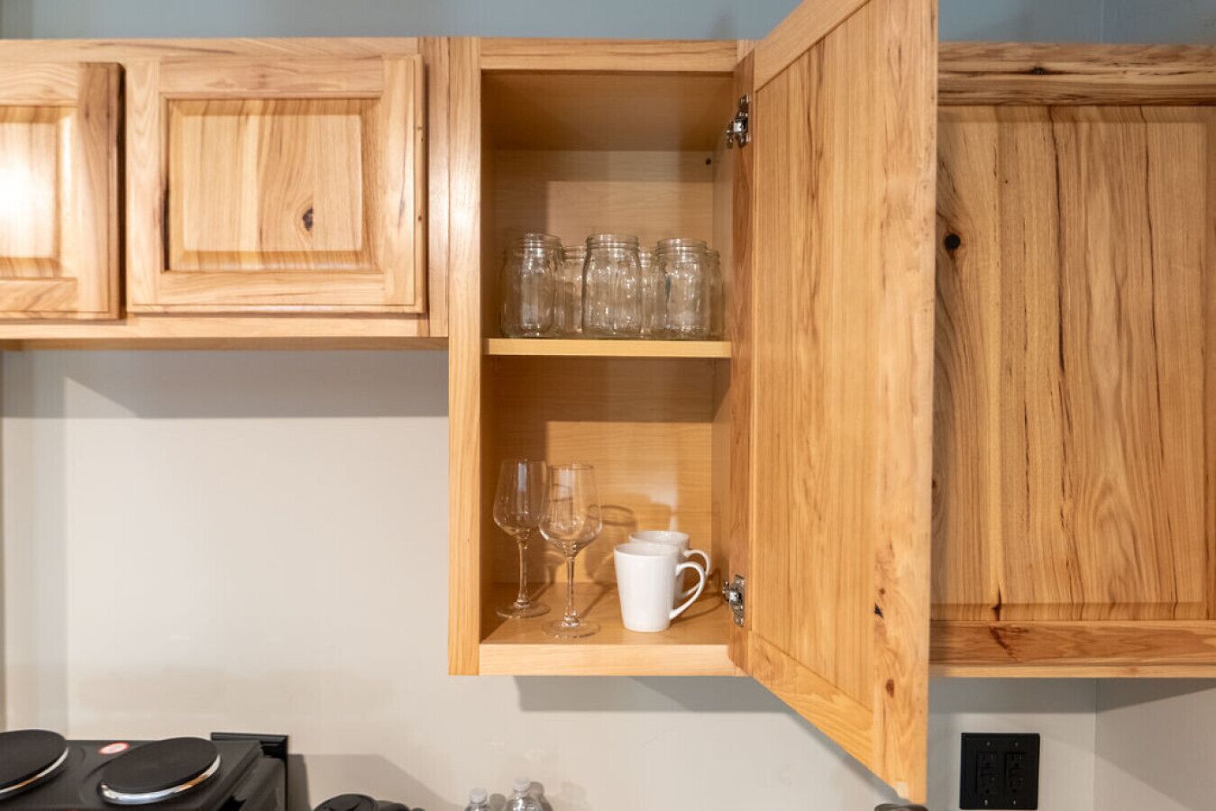 Un meuble en bois doté de deux étagères : l’une contient des bocaux en verre transparent, et l’autre, des verres à vin et une tasse blanche.
