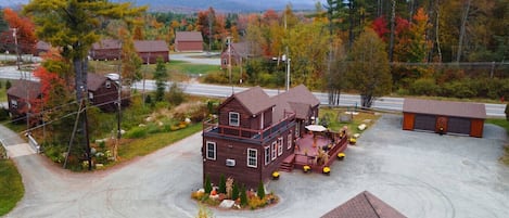 A two-story wooden house with a porch, surrounded by autumn foliage and a mountainous backdrop.