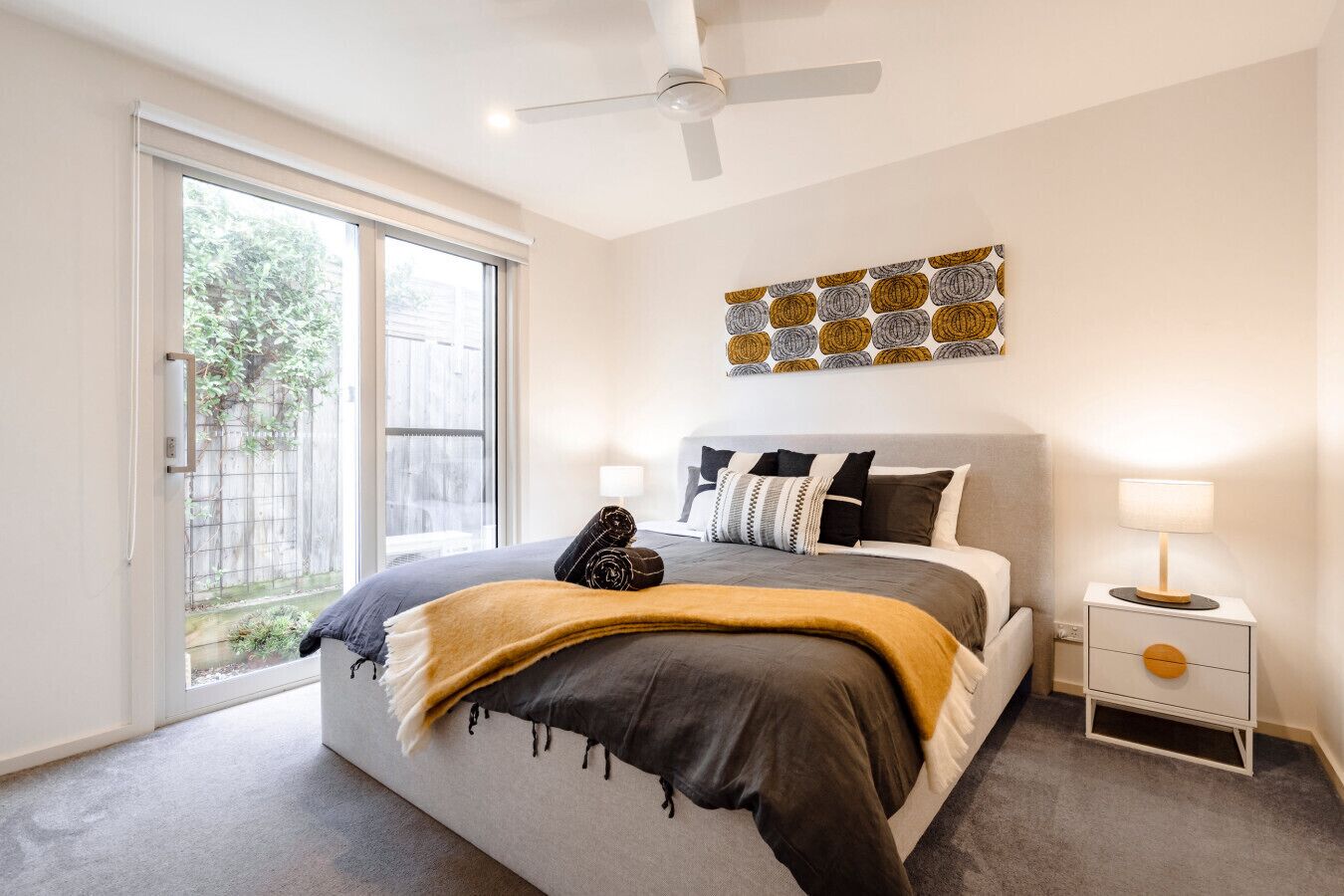 A bedroom with a bed, a ceiling fan, a nightstand with a lamp, and a window with a view of greenery.