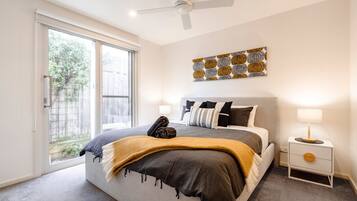 A bedroom with a bed, a ceiling fan, a nightstand with a lamp, and a window with a view of greenery.