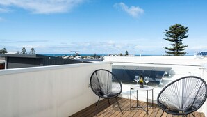 En tagterrasse med to sorte metalstole, et lille rundt bord og udsigt til havet og fjerne bygninger.