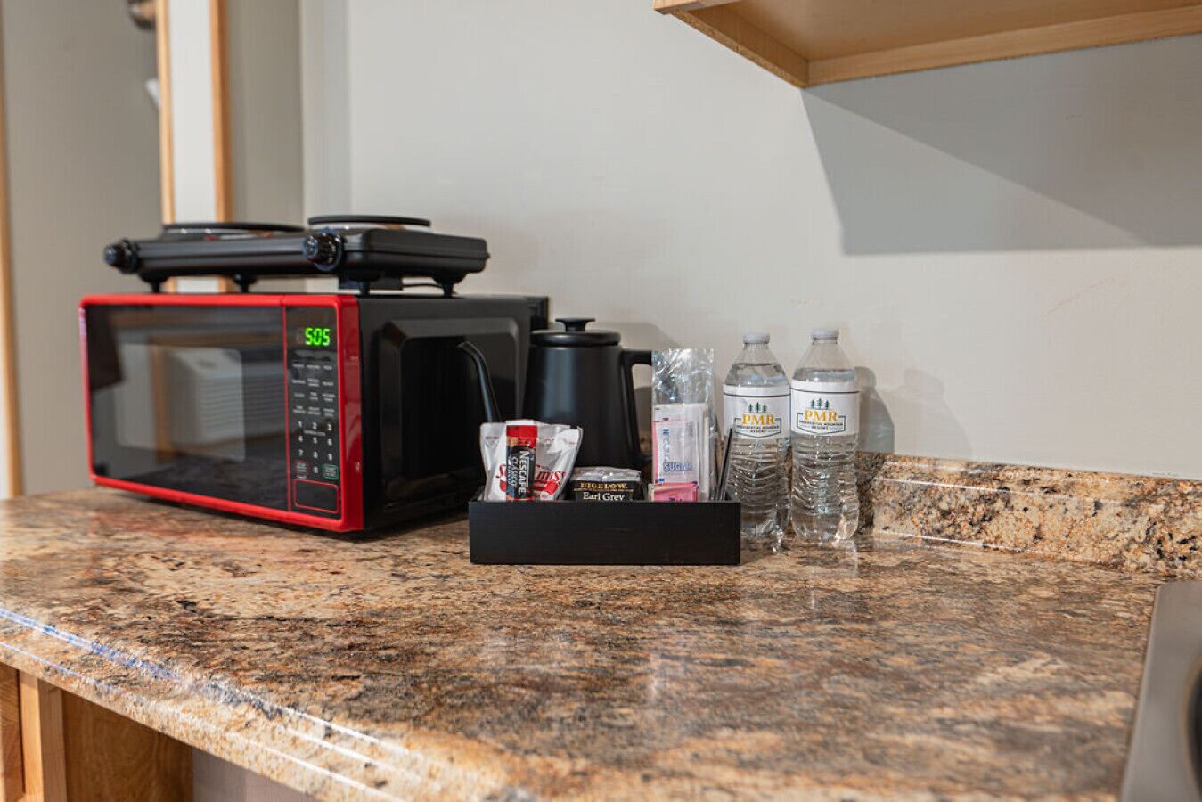 Un comptoir de cuisine avec un micro-ondes rouge et noir, une machine à café, des bouteilles d’eau et un plateau noir contenant des en-cas.