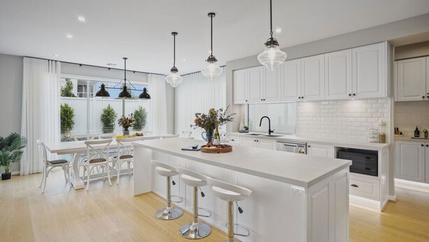 Una cocina moderna con armarios blancos, una isla central con taburetes y lámparas colgantes, y vistas al exterior a través de amplios ventanales.