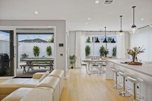 A modern kitchen with white countertops, bar stools, pendant lights, and a dining area with a table and chairs. - Hamptons in Torquay (499550) (Torquay)