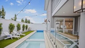 Un área de piscina exterior moderna con una piscina azul cristalina, sillas de descanso blancas y un césped cuidado.