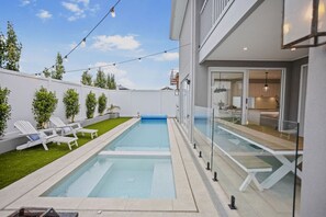 A modern outdoor swimming pool with lounge chairs, a grassy area, and a building with a glass door. - Hamptons in Torquay (499550) (Torquay)