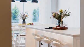 Una cocina moderna con taburetes blancos, una isla central con un florero y un comedor con sillas blancas y una mesa.