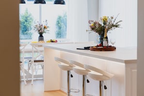 A modern kitchen with white bar stools, a dining table with chairs, and a window with a view of greenery. - Hamptons in Torquay (499550) (Torquay)