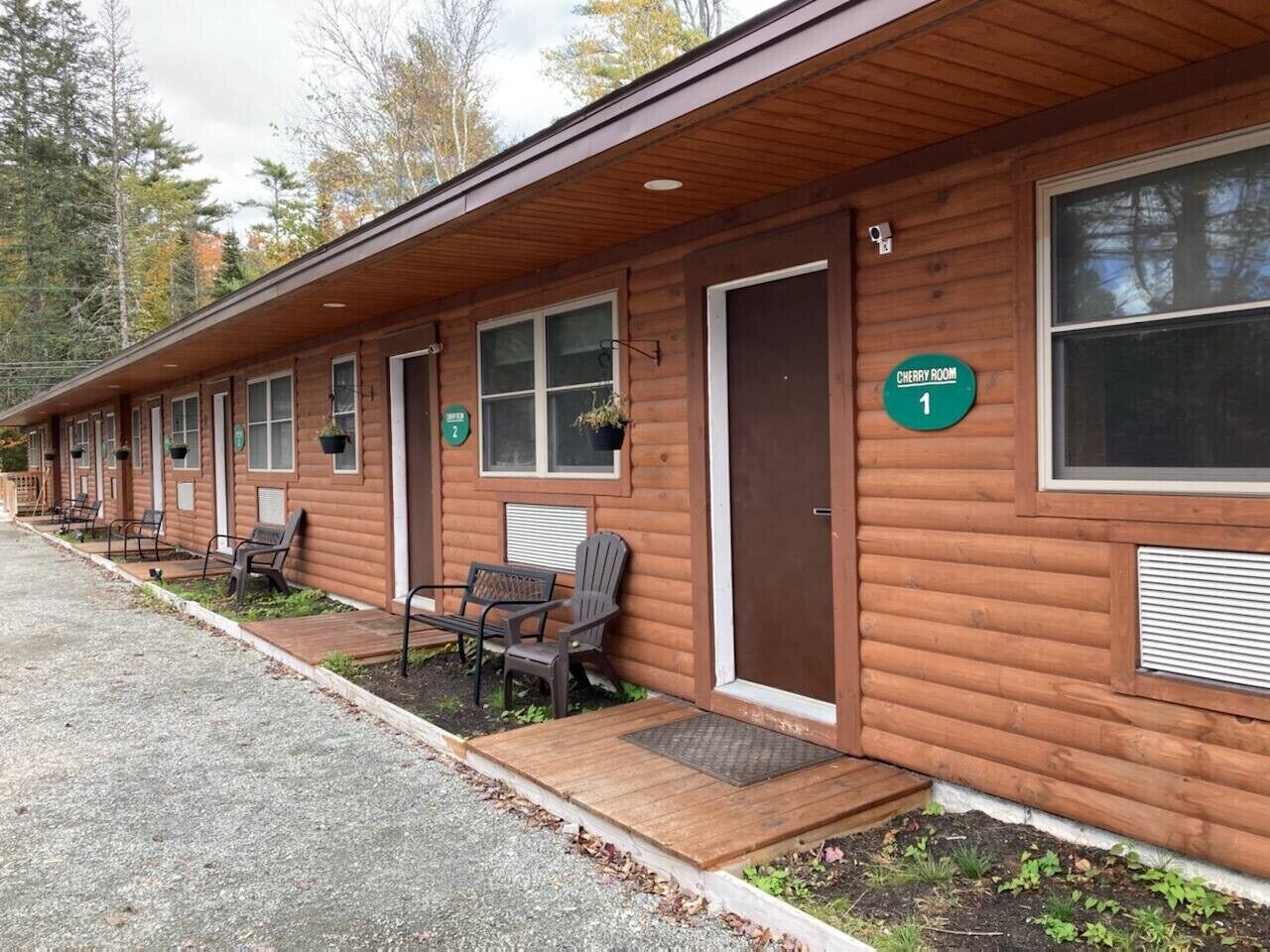 A row of log cabins with numbered doors, outdoor seating, and a sign indicating 'Cherry Room 1'.