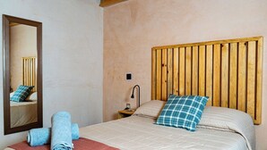 Habitación de hotel con cabecera de madera, cama con colcha color beige, dos almohadas, un espejo, una mesita de noche con lámpara y toallas enrolladas sobre la cama.