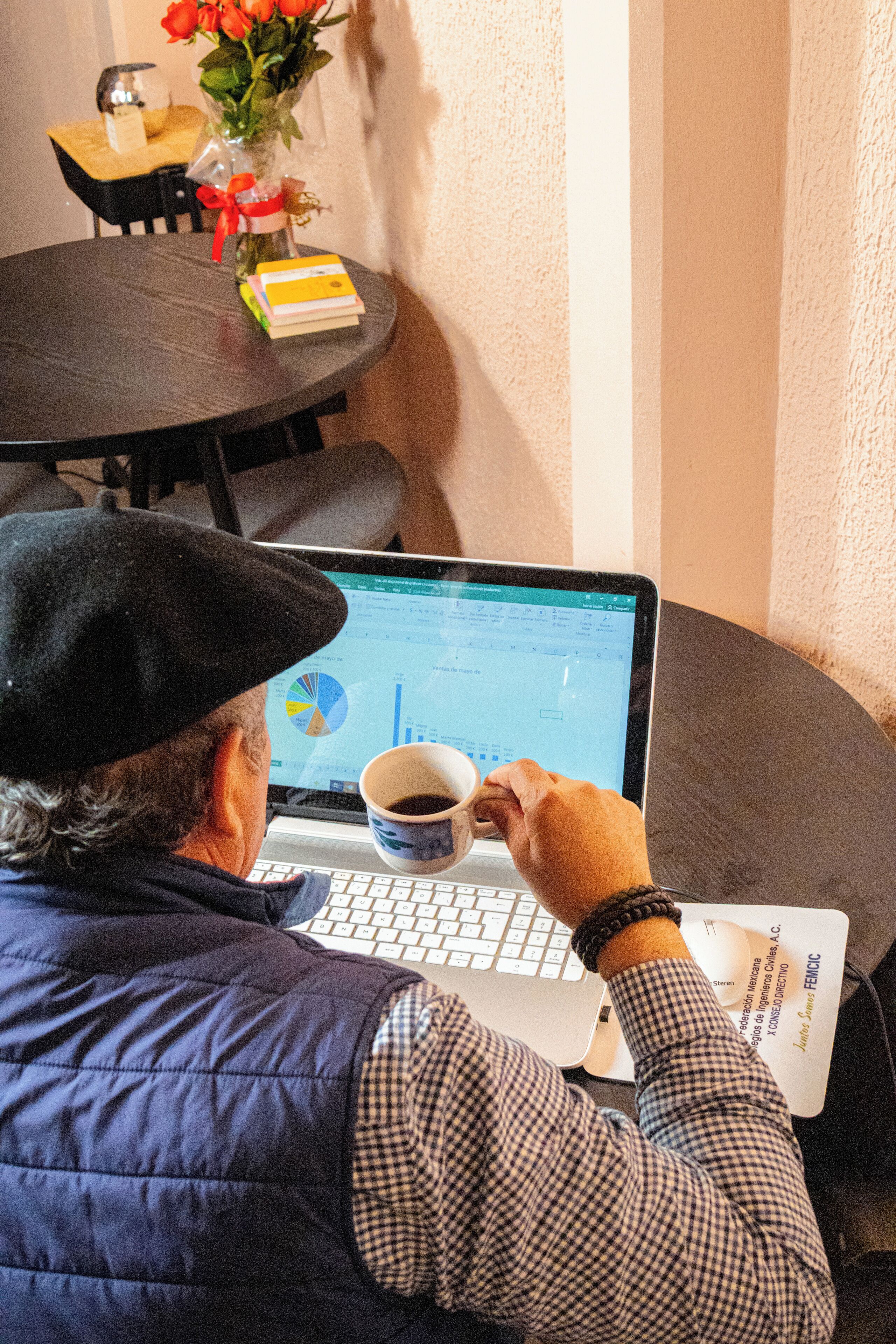 A person using a laptop at a round table with a cup of coffee, a book, and a vase with flowers.
