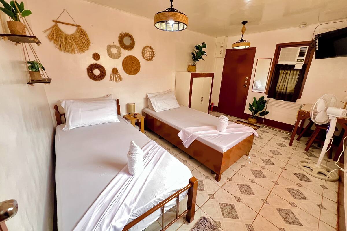 A bedroom with two beds, a wooden wardrobe, a desk with a chair, a hanging light fixture, and wall decorations.