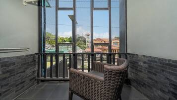 Uma varanda com cadeira de vime, guarda-corpo de metal e vista para edifícios e vegetação.