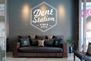 A dark-colored leather sofa with multiple cushions, a logo on the back wall, and a potted plant to the right. - Dent Station Residence (Chalong)