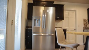 A kitchen with a stainless steel refrigerator, dark cabinets, a light-colored chair, and a tiled floor. - Cozy Stylish 2 BR Home w / Relax & Enjoy (Owensboro)