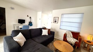 Een woonkamer met een hoekbank, een klein rond tafeltje, een rode fauteuil, een televisie, een gitaar en een raam met jaloezieën.